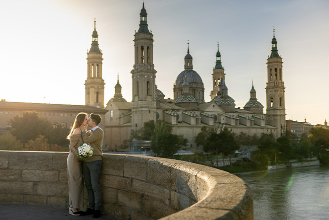 Yaiza y Salomón en su preboda en Zaragoza, abrazados con un ramo de flores frente a la Basílica del Pilar iluminada por el atardecer.